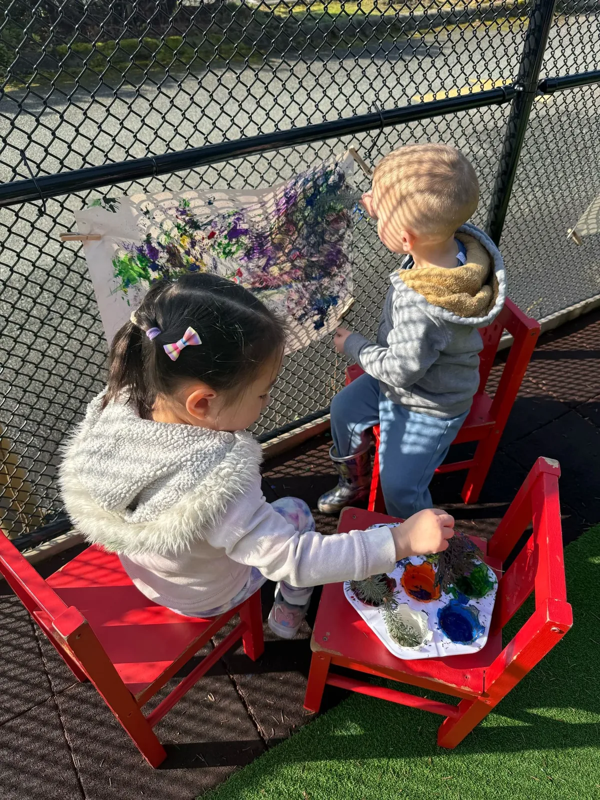 Two children painting on a large canvas clipped to a fence during outdoor art time