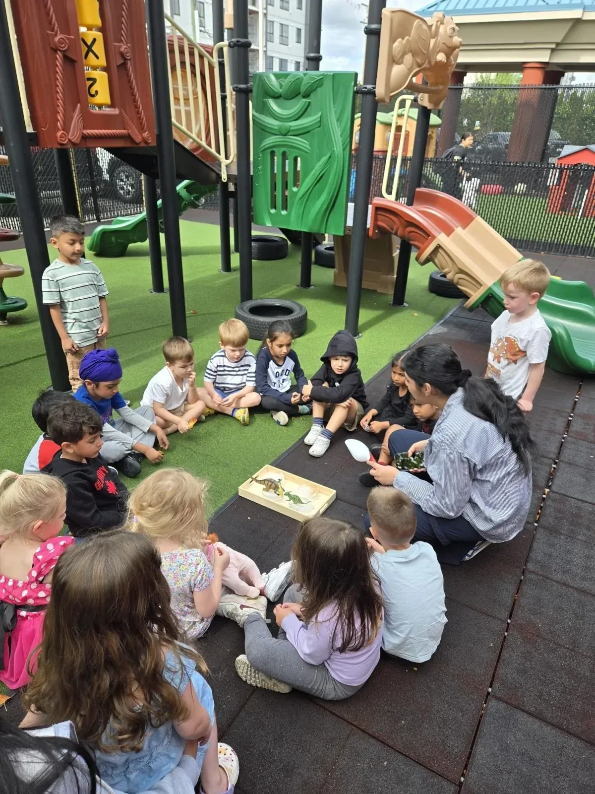 An educator and a group of children gathered in a circle on the playground exploring a discovery tray