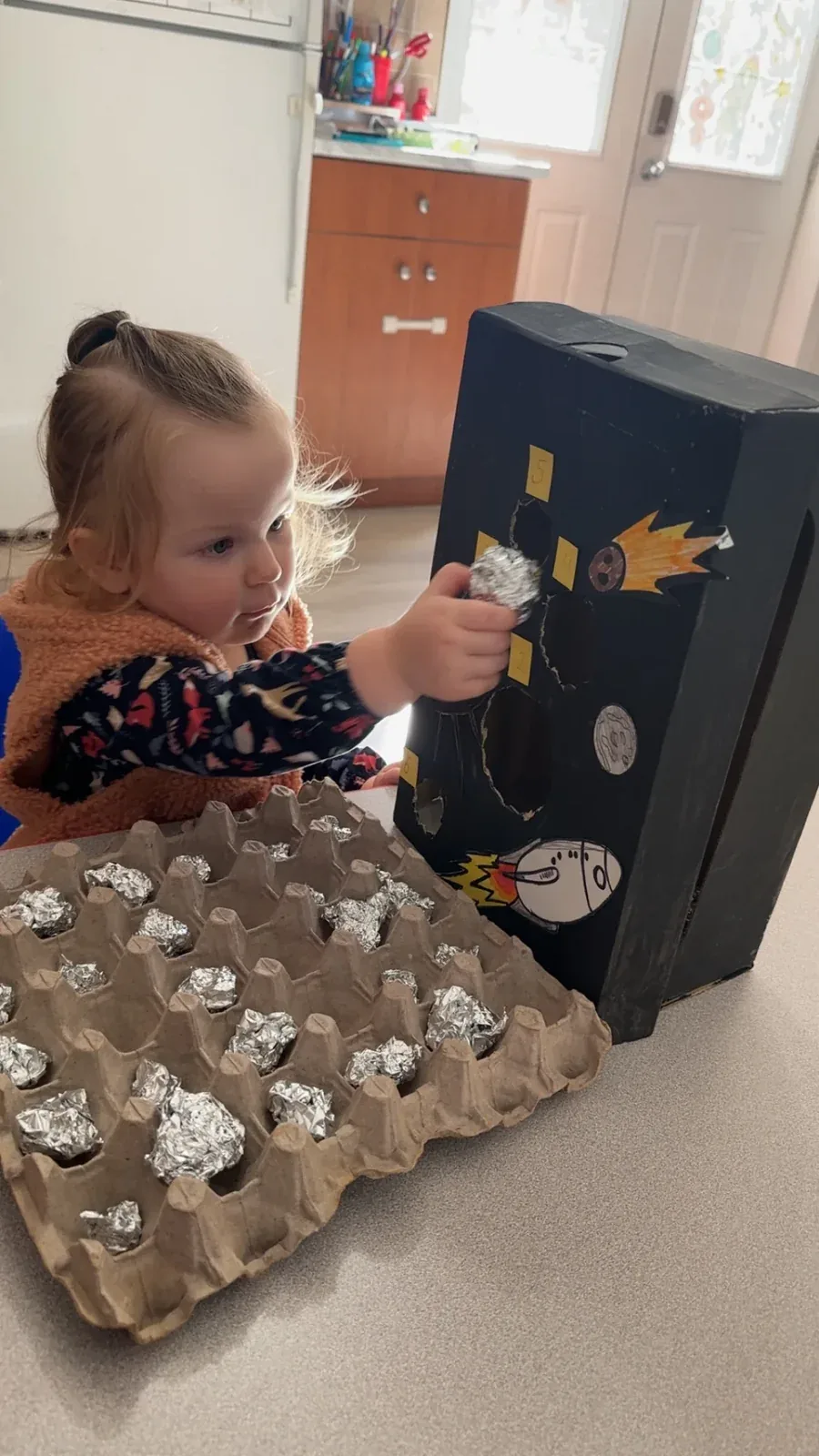 A toddler placing foil pieces into a handmade cardboard rocket craft