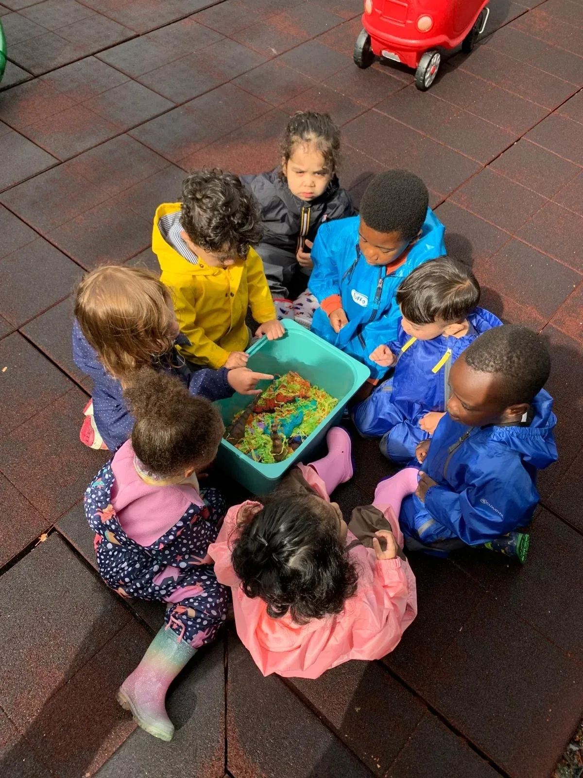 A group of children in rain jackets gathered around a colourful sensory bin outdoors