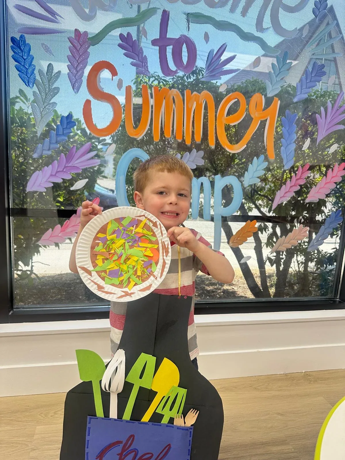 A smiling summer camper in a chef apron holding a paper-plate craft in front of the Welcome to Summer Camp mural