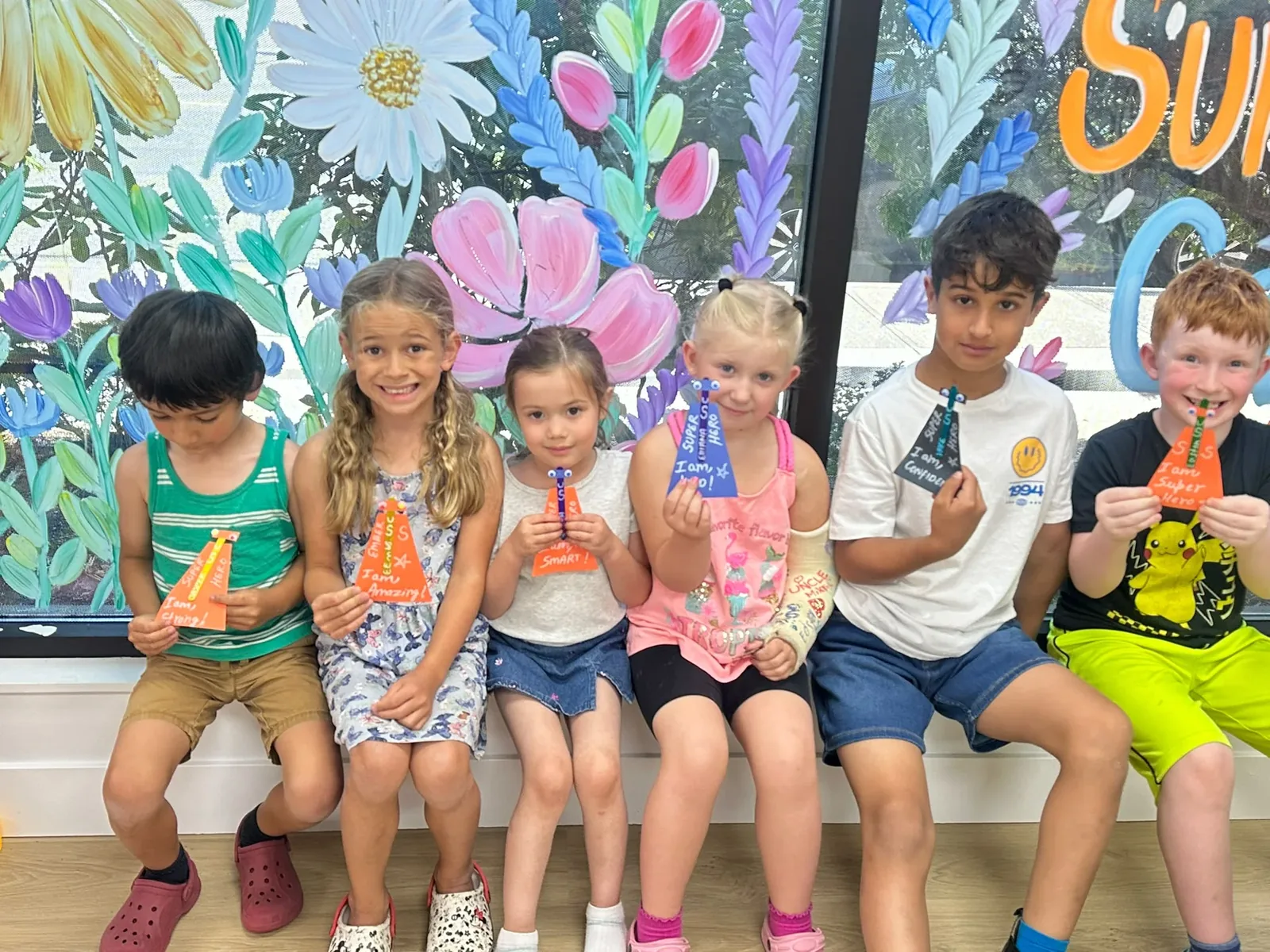 Summer camp children holding their rocket handprint art in front of a floral Welcome to Summer Camp mural