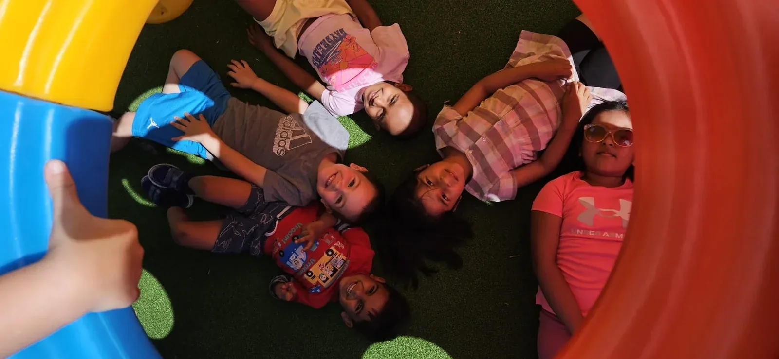 Children lying in a circle on turf, smiling up through a play tunnel