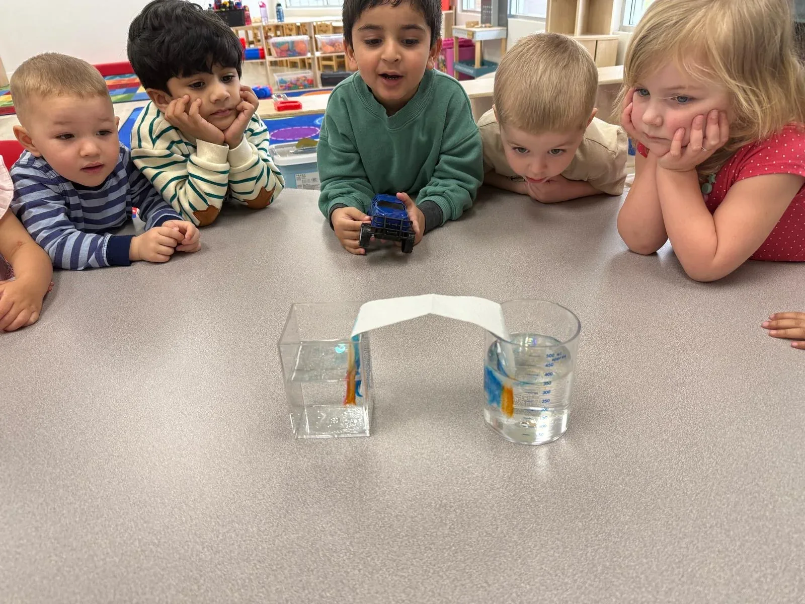 Children watching a capillary-action water experiment with two beakers and a paper bridge